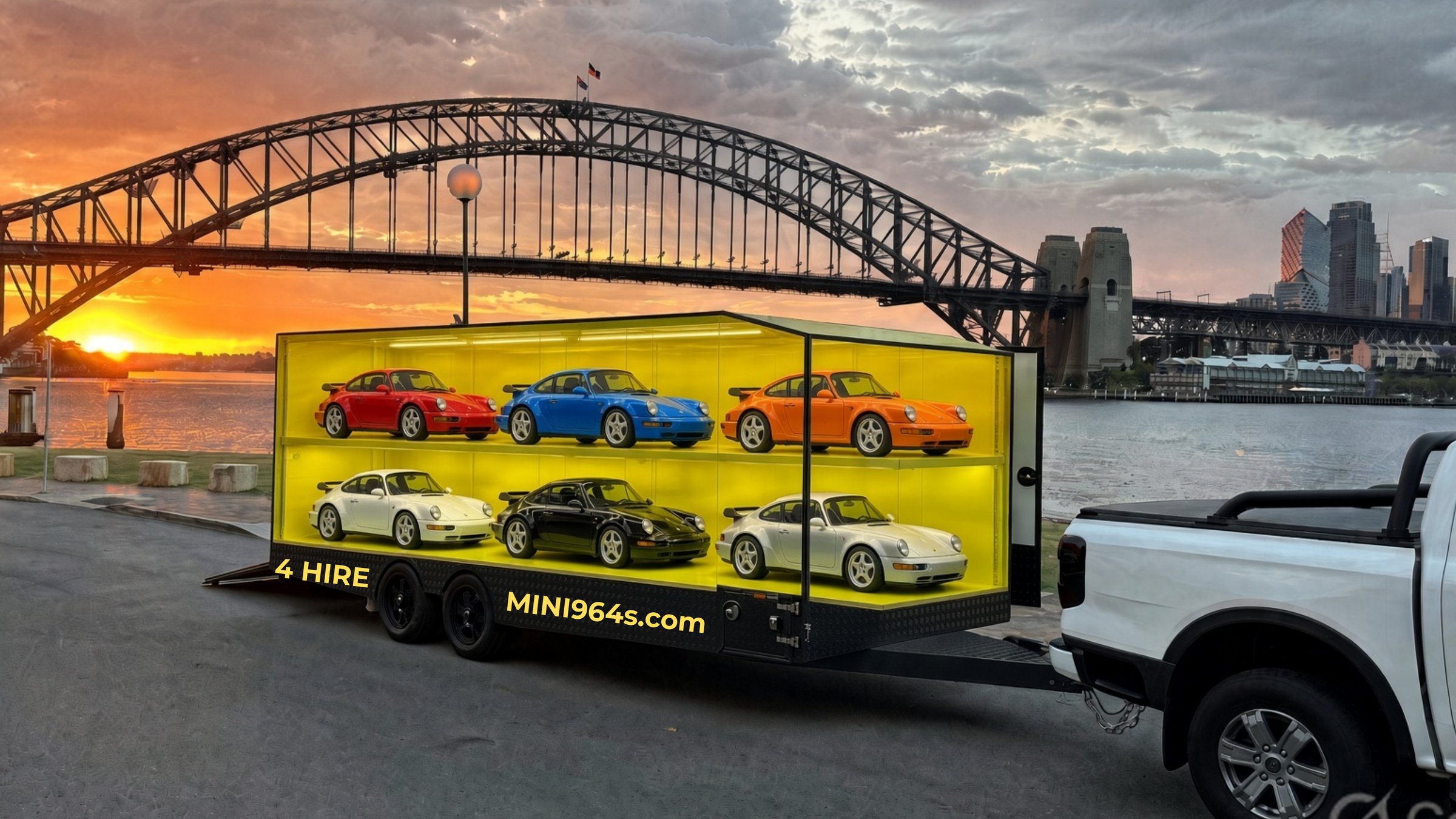 Micro Bespoke Garage display trailer — yellow perspex showcase with Junior cars, underglow lighting, parked at Sydney Harbour Bridge sunset